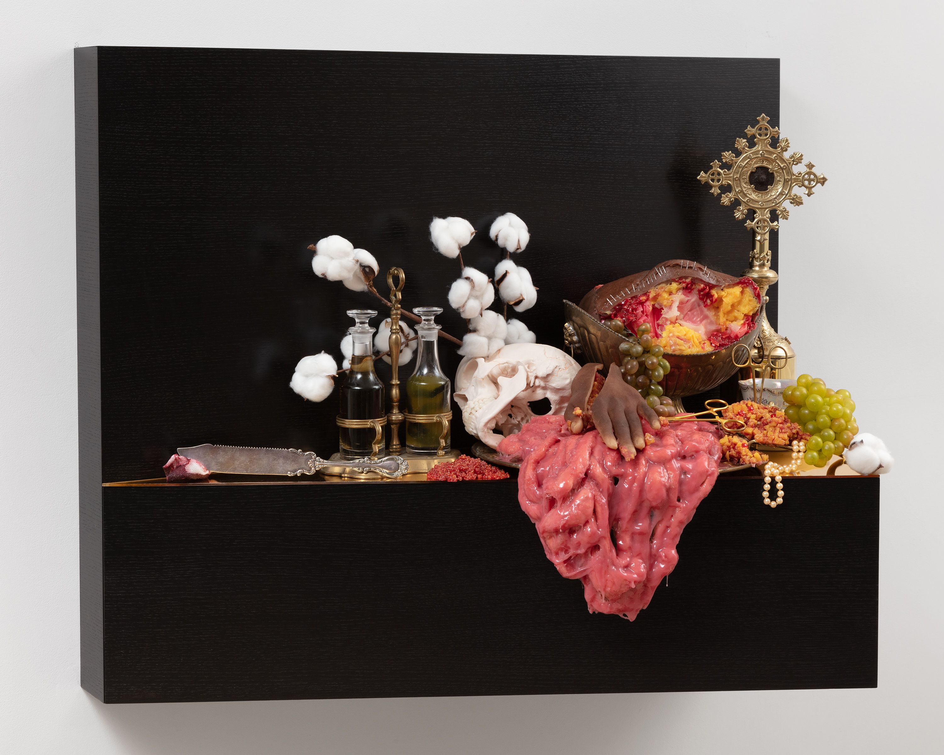 This sculpture is a wooden shelf mounted to wall. The shelf is stained a very dark brown, and is covered in a number of objects, including a severed hand, flesh draped to the side, grapes, branches of cotton, scissors, an antique silver knife, and a pearl necklace.