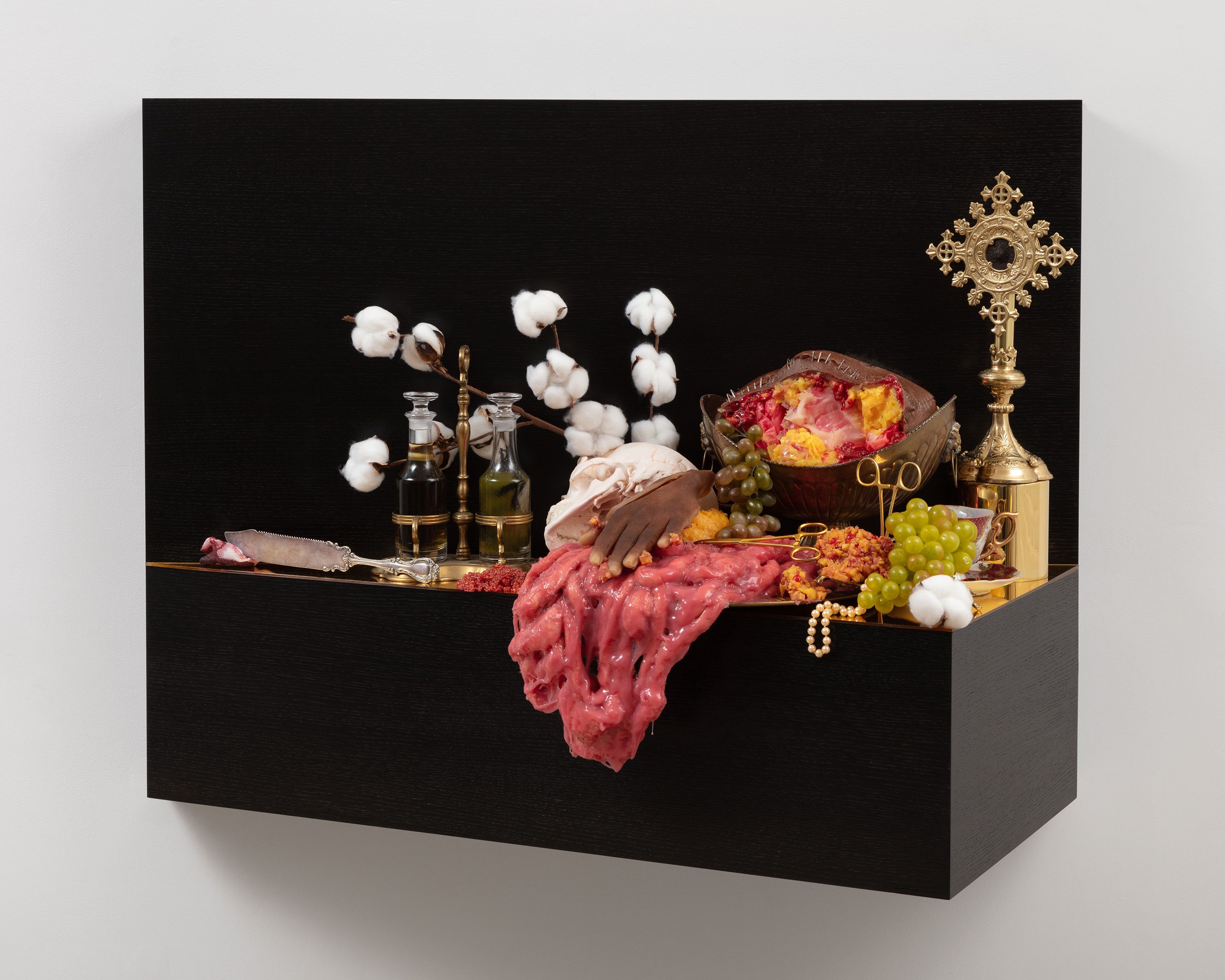 This sculpture is a wooden shelf mounted to wall. The shelf is stained a very dark brown, and is covered in a number of objects, including a severed hand, flesh draped to the side, grapes, branches of cotton, scissors, an antique silver knife, and a pearl necklace.